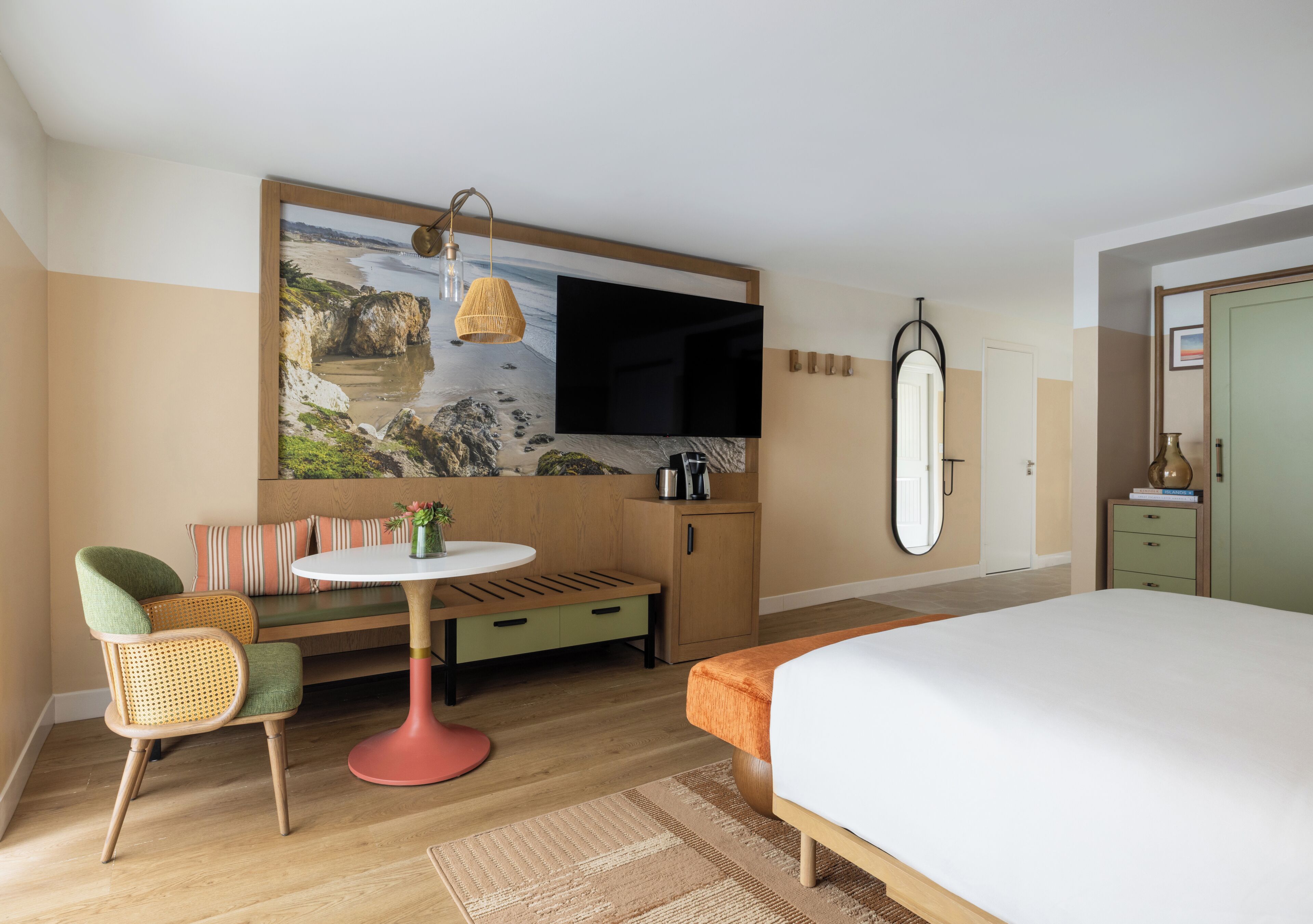 Habitación de hotel con cama, zona de estar con mesa, televisión y un cuadro grande de una playa.