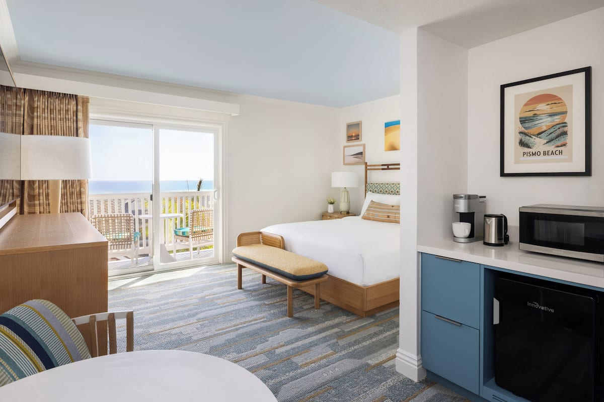 A hotel room with a bed, a desk, a bench, a kitchenette, and a large window with a view of the beach.