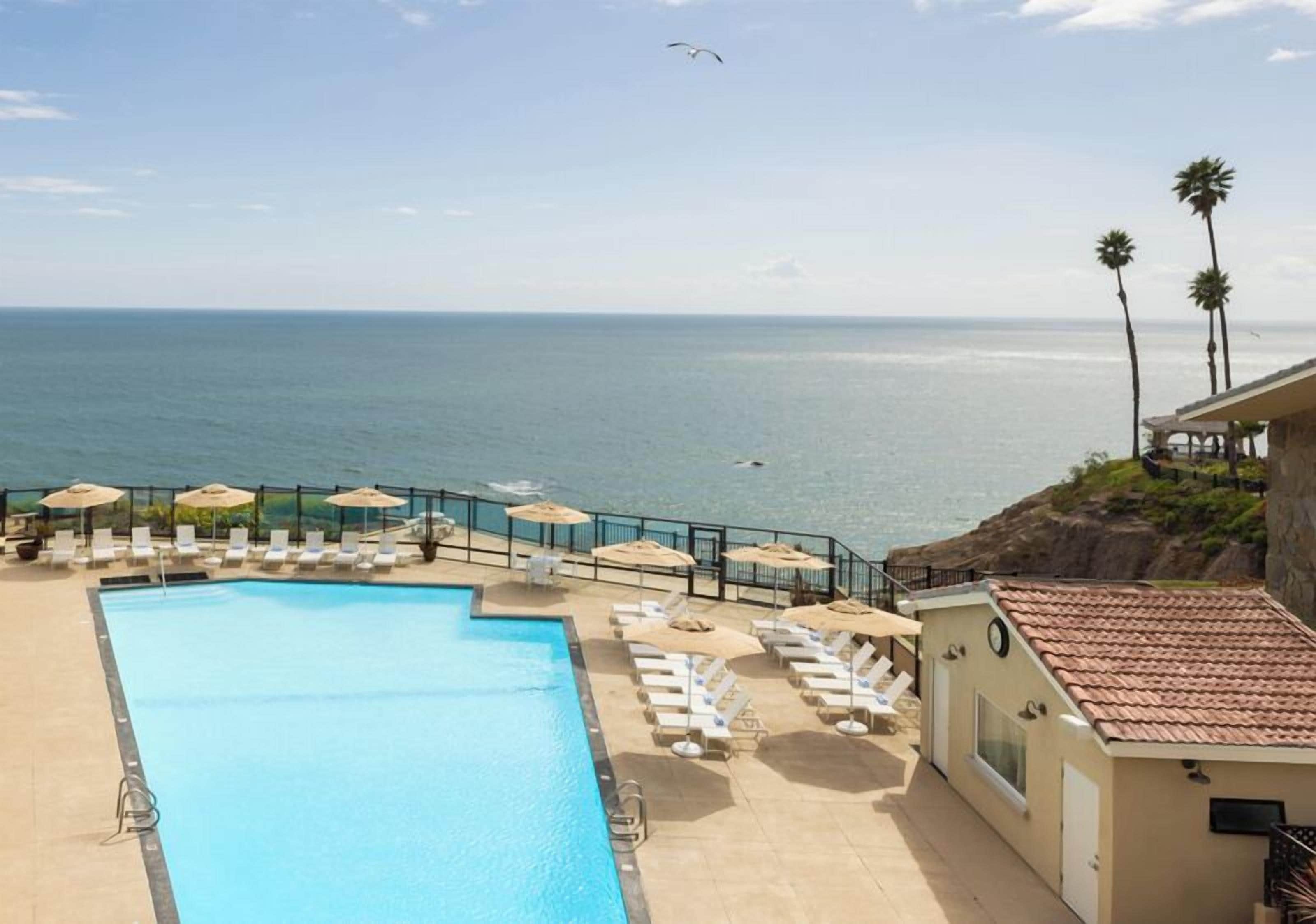 A pool with lounge chairs and umbrellas, overlooking a beach and ocean.