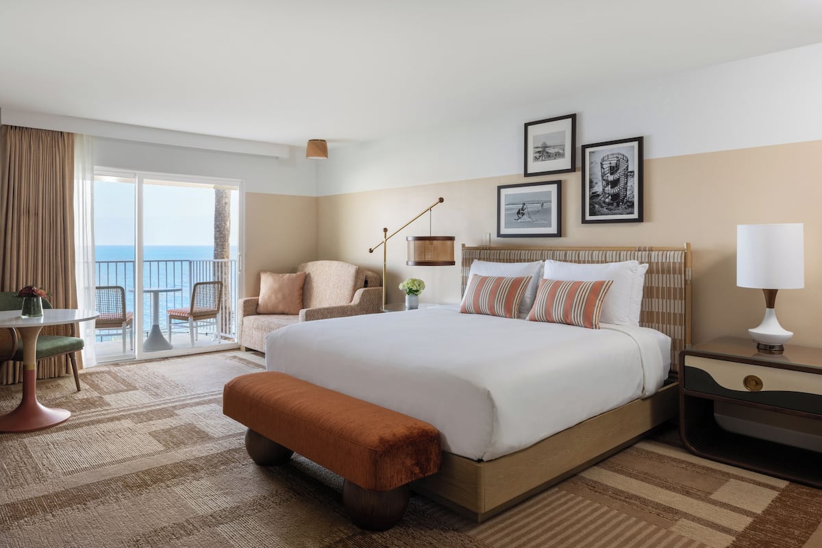 A hotel room with a large bed, a desk, a chair, a bench, a lamp, and a view of the sea.