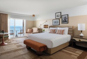 Habitación de hotel con una cama grande, un escritorio, una silla, un banco, una lámpara y vistas al mar.