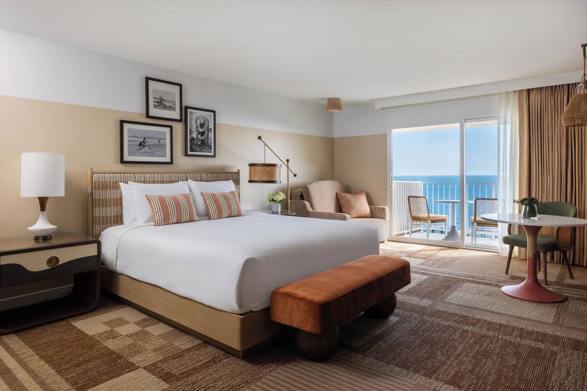 A hotel room with a large bed, a bedside table, a bench, a chair, a small table, and a balcony with a view.