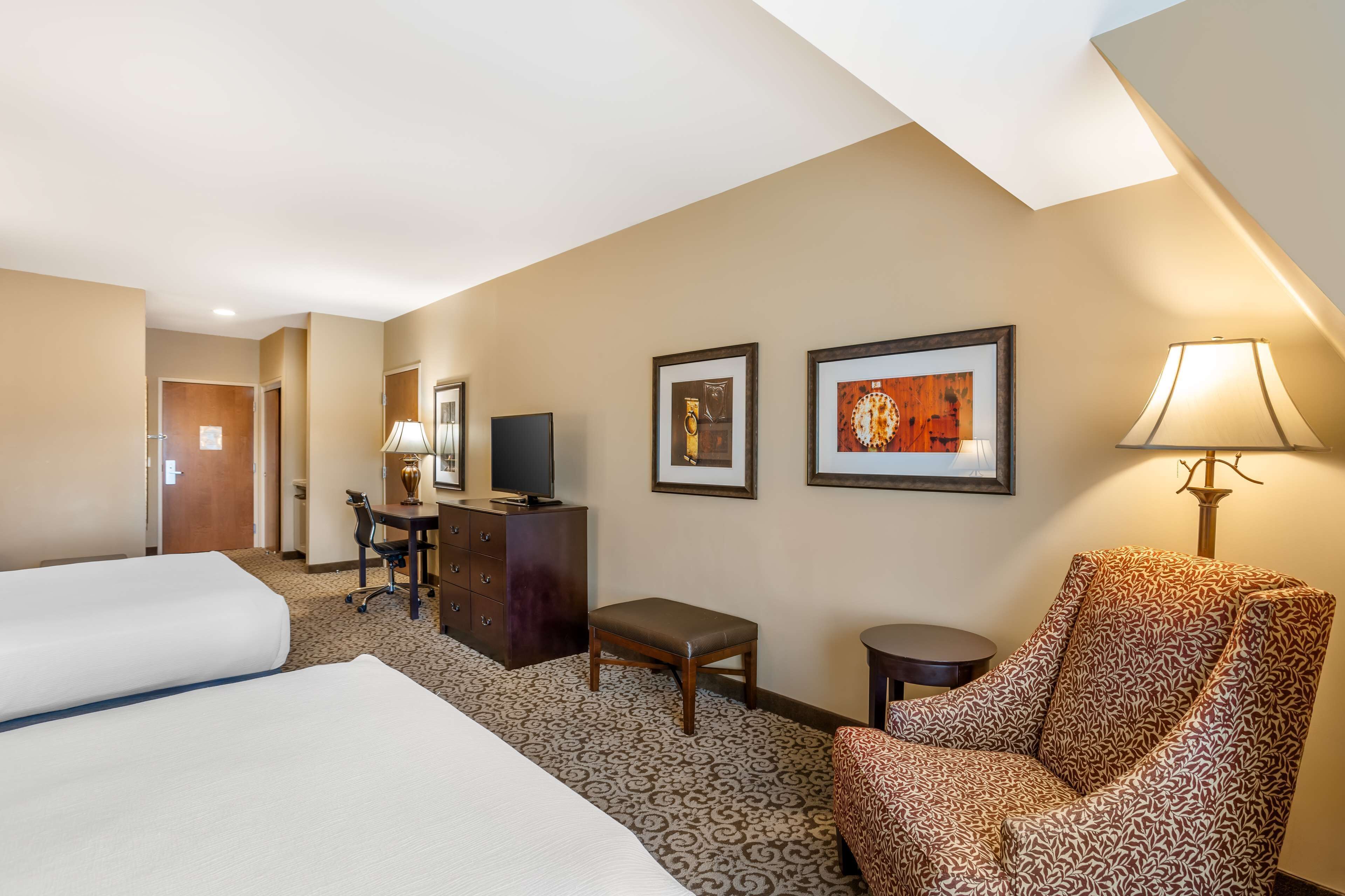 A hotel room with two beds, a desk with a chair, a small table, a lamp, and two framed pictures on the wall.