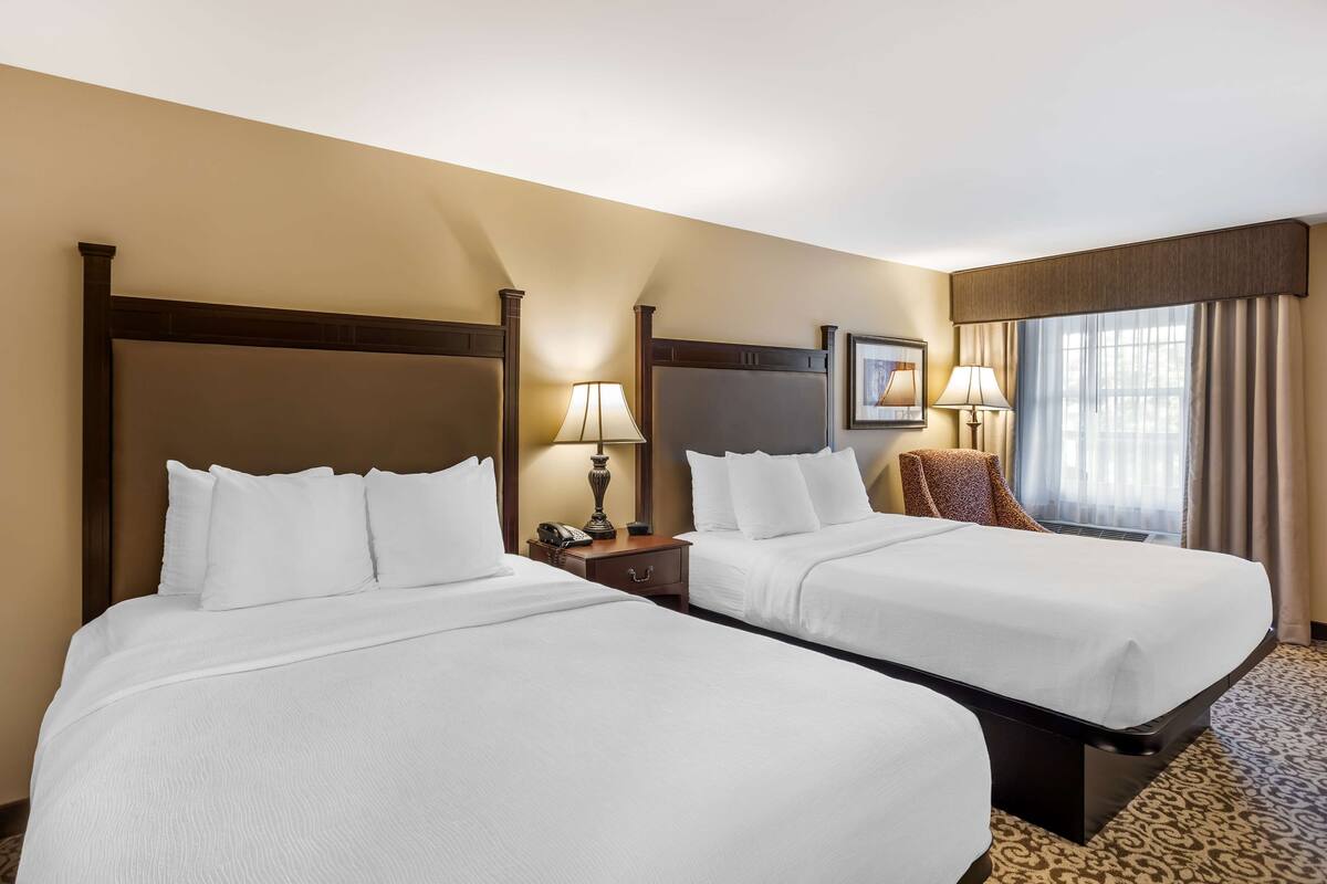 A hotel room with two beds, a nightstand with a lamp, a chair, and a window with curtains.