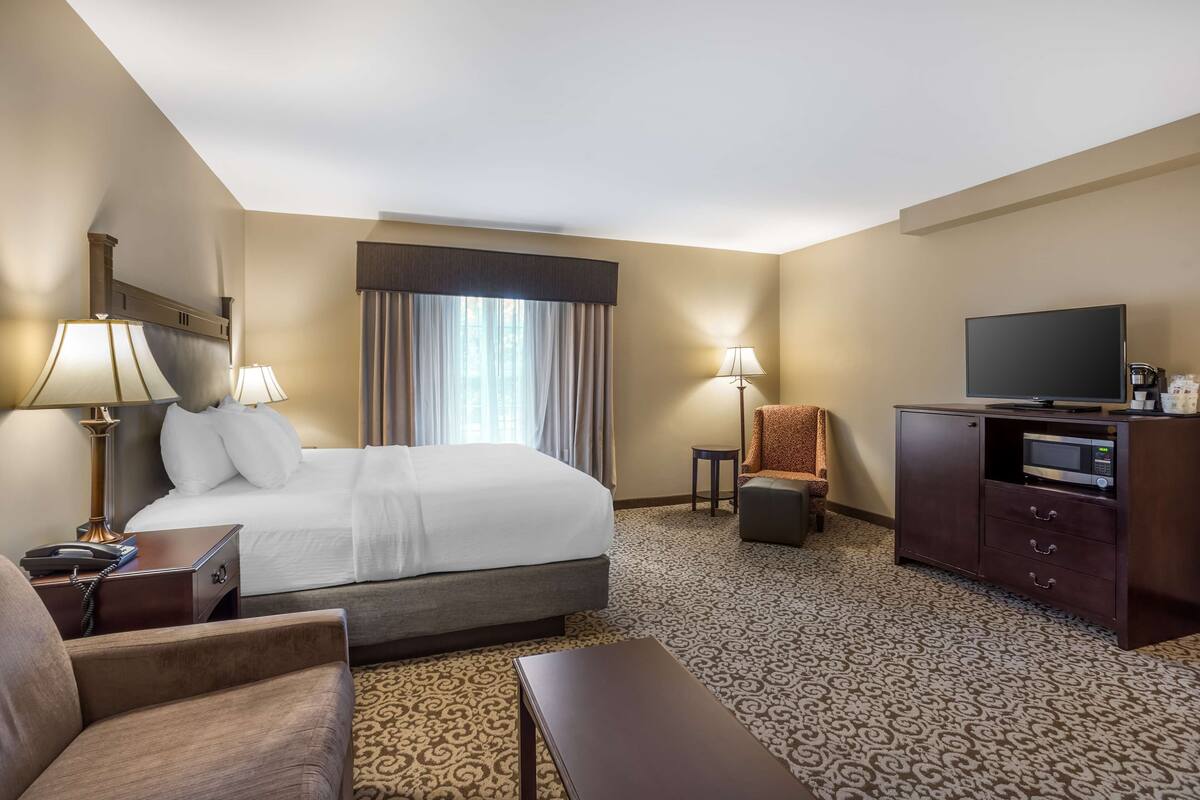 A hotel room with a large bed, two bedside lamps, a flat-screen TV, a small table, a chair, and a cabinet with a microwave.