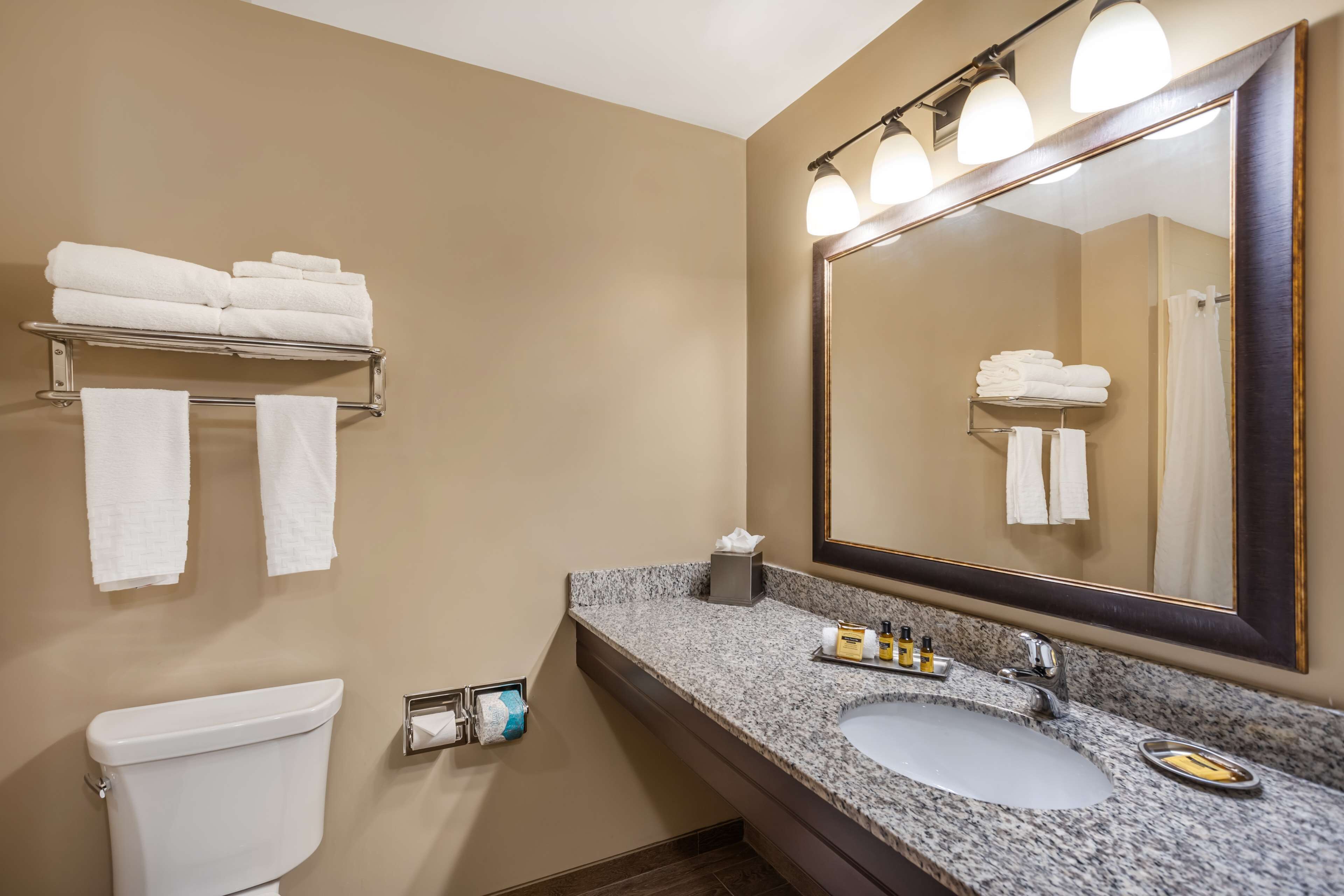 A bathroom with a toilet, sink, and mirror. There are towels on a rack and a shelf, and toiletries on the sink counter.