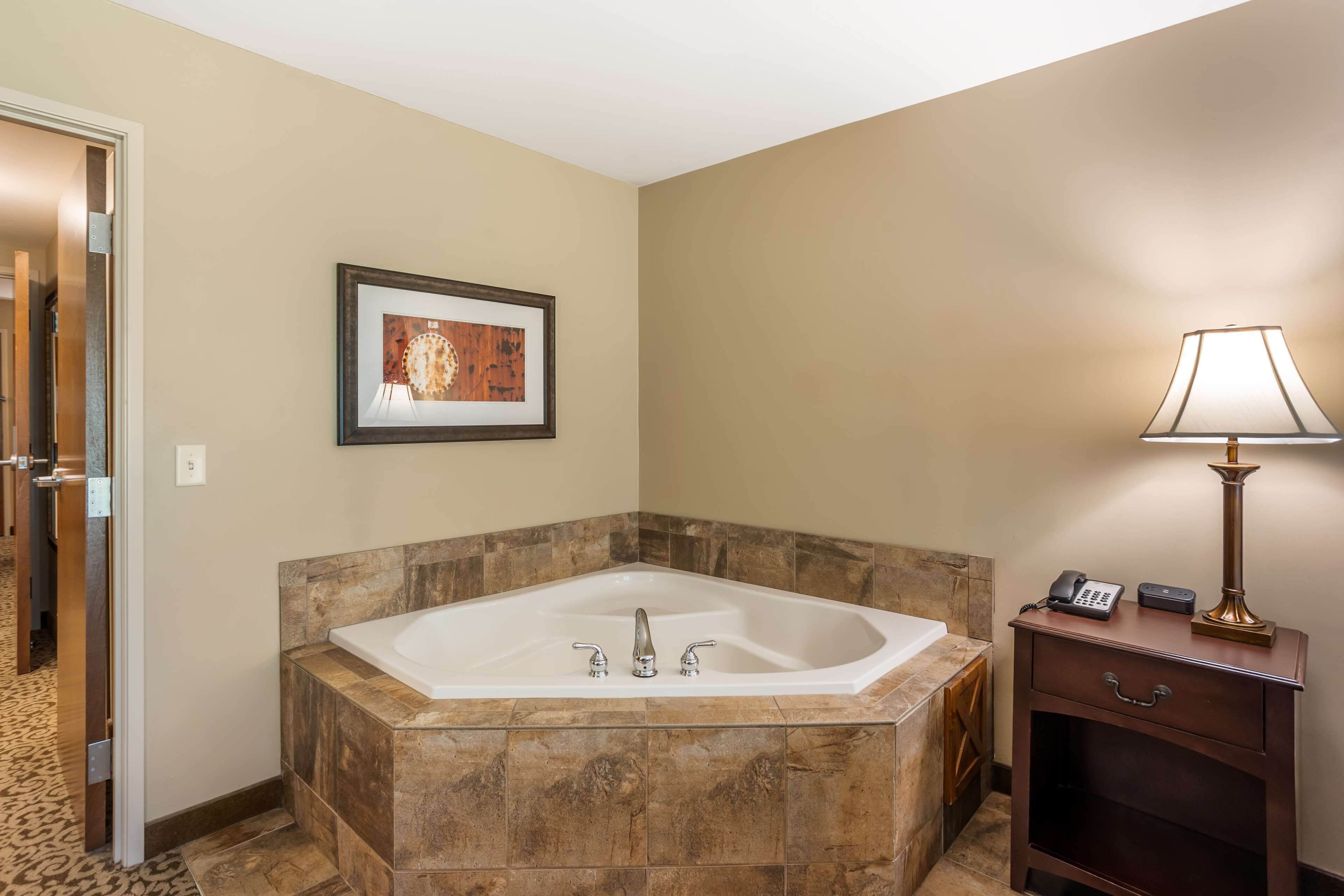 A bathroom with a large freestanding bathtub, a framed artwork on the wall, a bedside table with a lamp, and a door leading to another room.