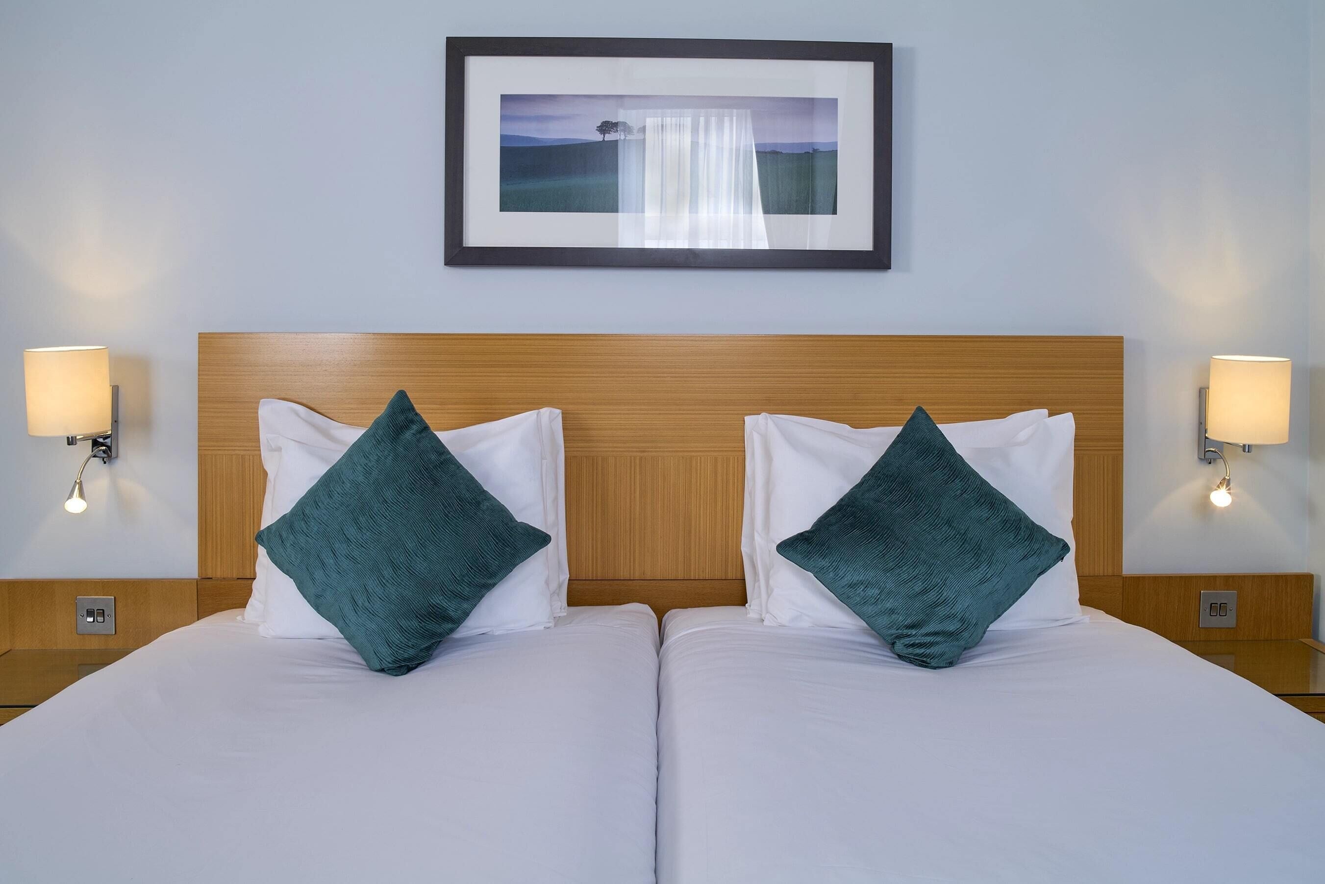 A hotel room with two beds, each with white bedding and teal pillows, a wooden headboard, and a framed picture on the wall.
