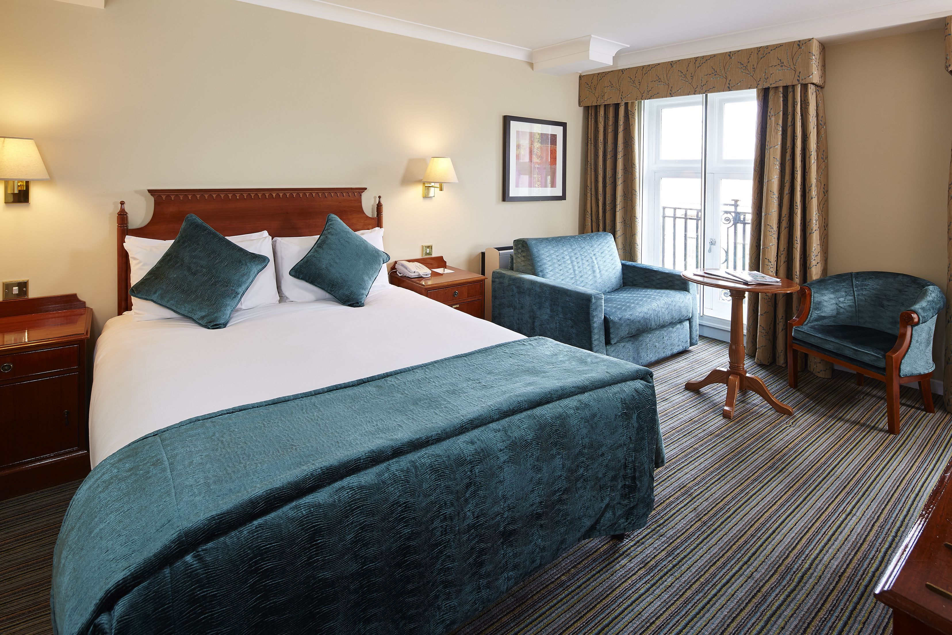 A hotel room with a large bed, two armchairs, a small table, and a view of the outdoors through a window.