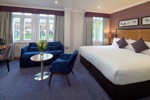 Ein Hotelzimmer mit einem großen Bett, zwei blauen Sesseln, einem kleinen runden Tisch mit Blumen und Blick durch große Fenster nach draußen.