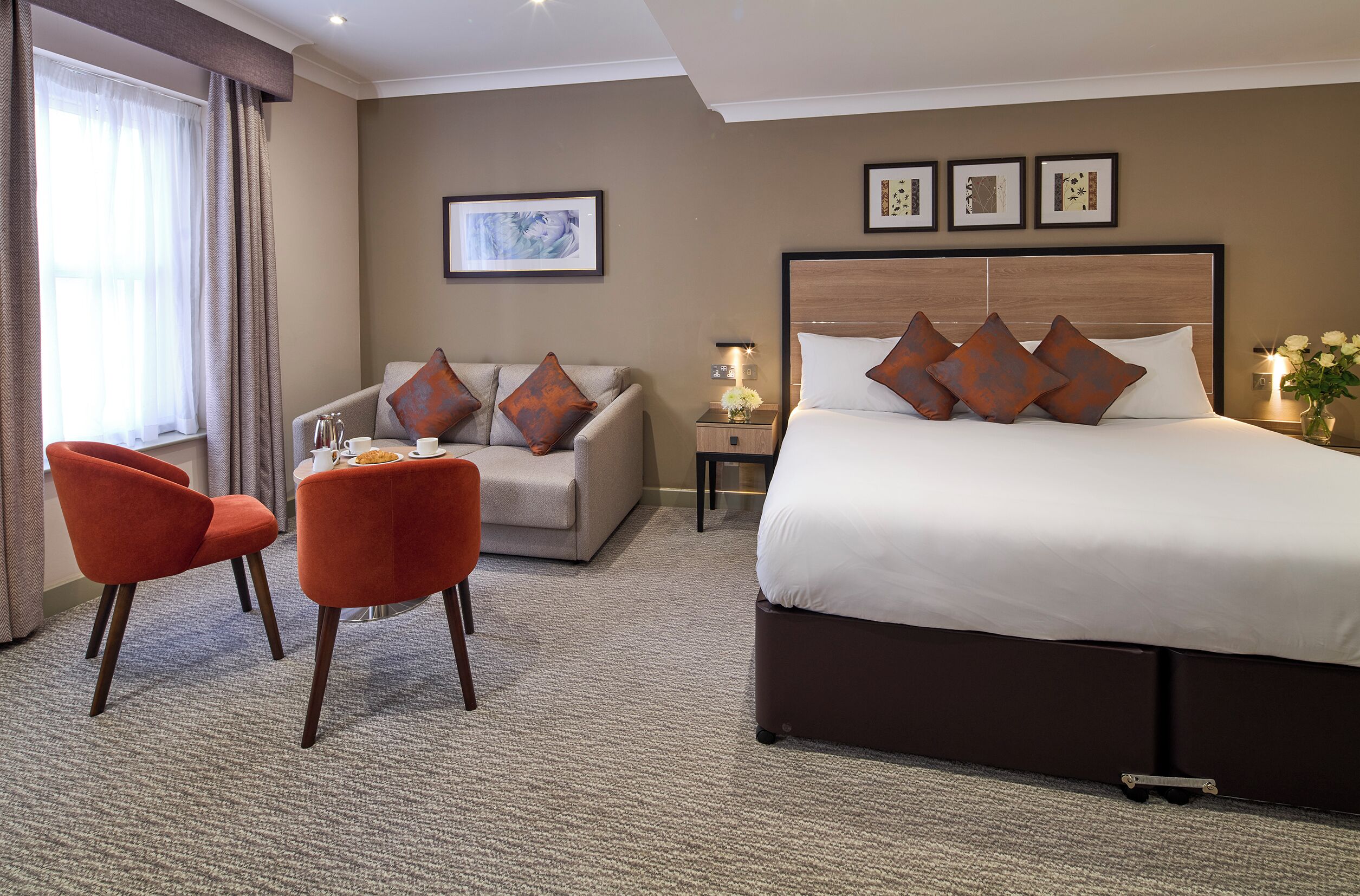 A hotel room with a large bed, a sofa, two armchairs, a small table with a vase of flowers, and framed artwork on the wall.