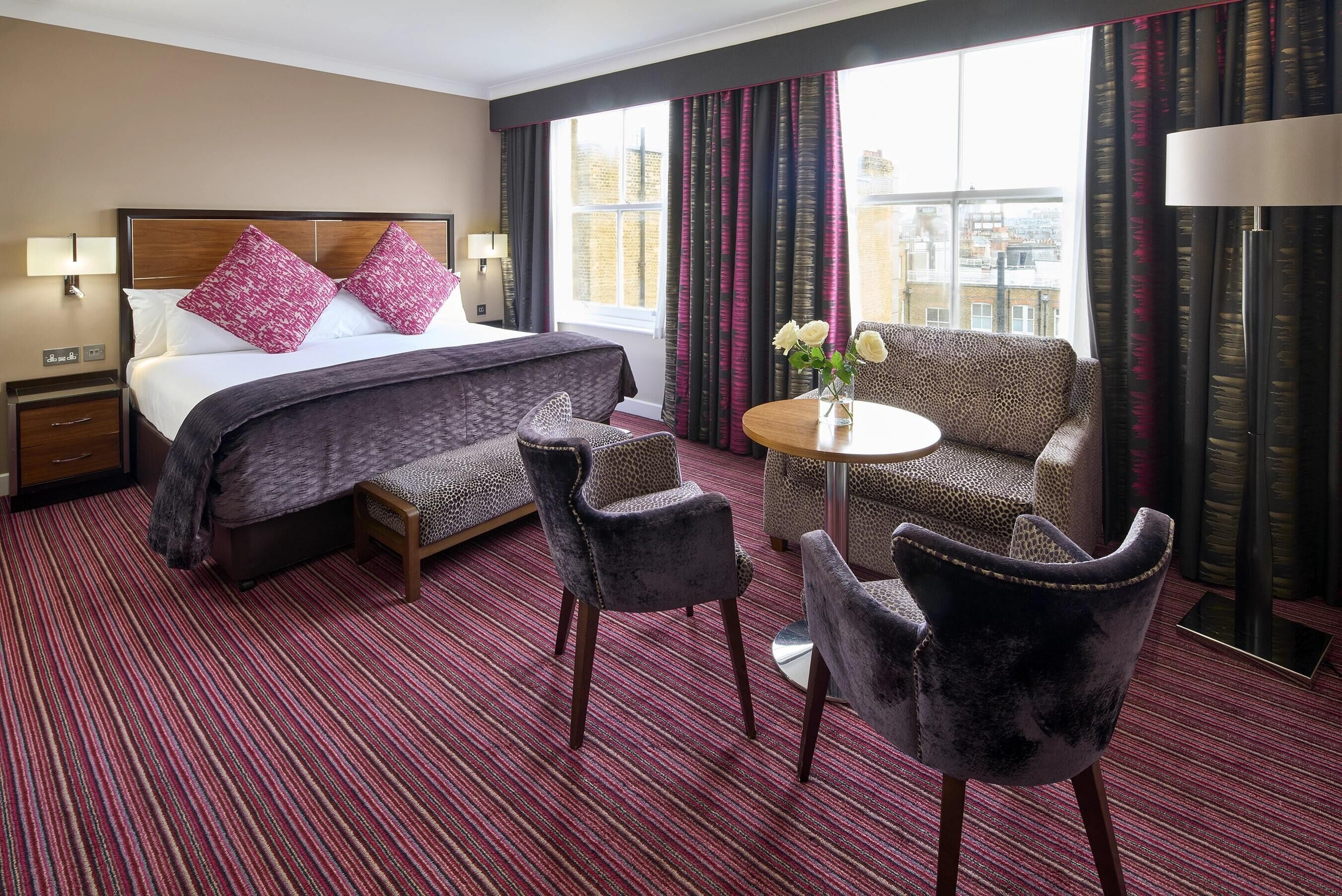 Ein Hotelzimmer mit einem großen Bett, zwei Sesseln, einem kleinen Tisch und Blick auf die Stadt durch das Fenster.