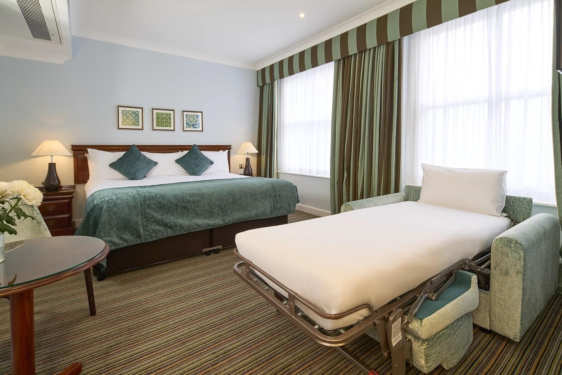 Quarto de hotel com cama, mesa de cabeceira, cadeira e uma pequena mesa.