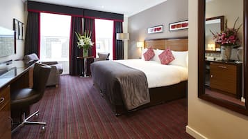 A hotel room with a large bed, a desk, a chair, a desk lamp, a mirror, and a window with curtains.