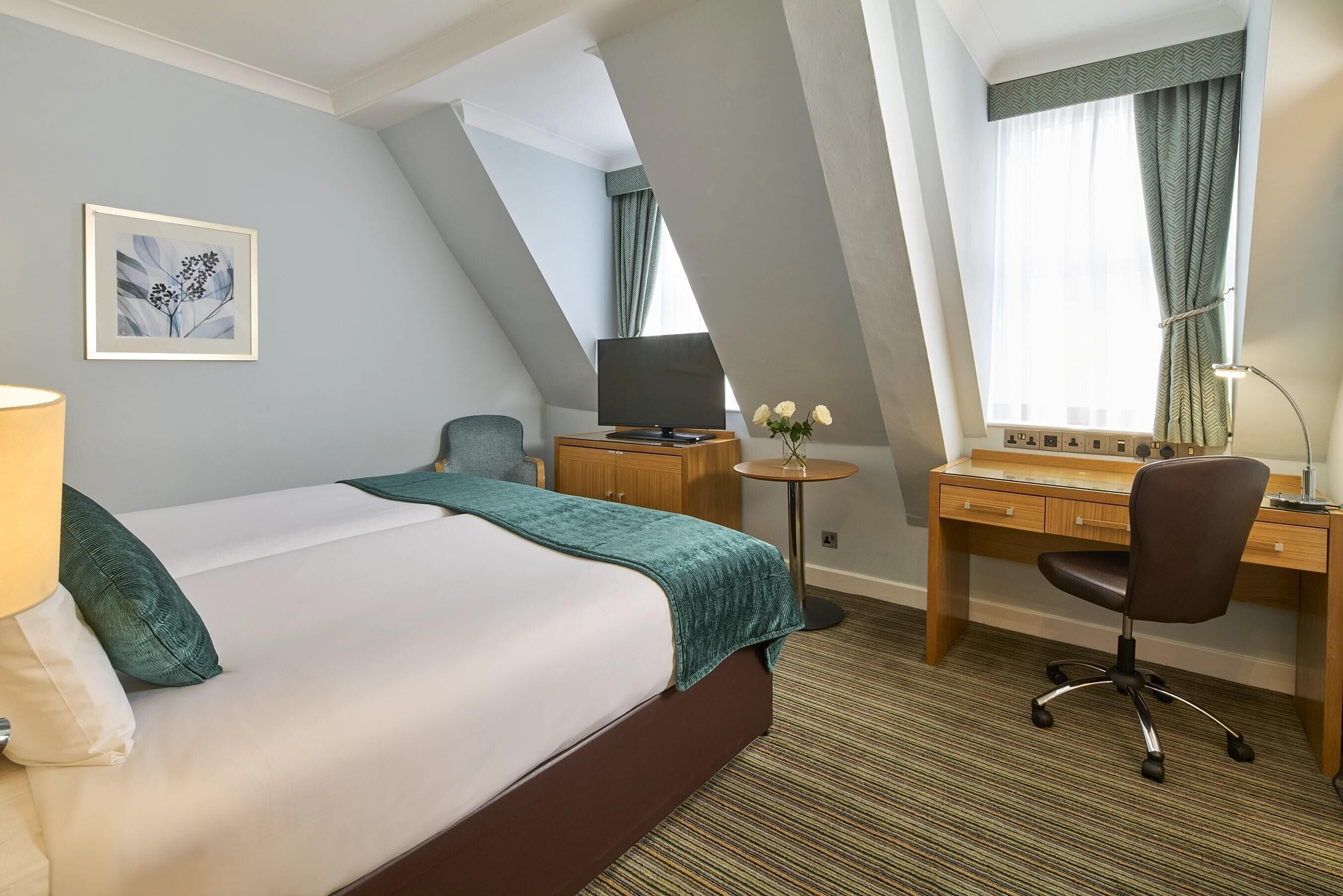 A hotel room with a bed, a desk, a chair, a television, and a small table with flowers.