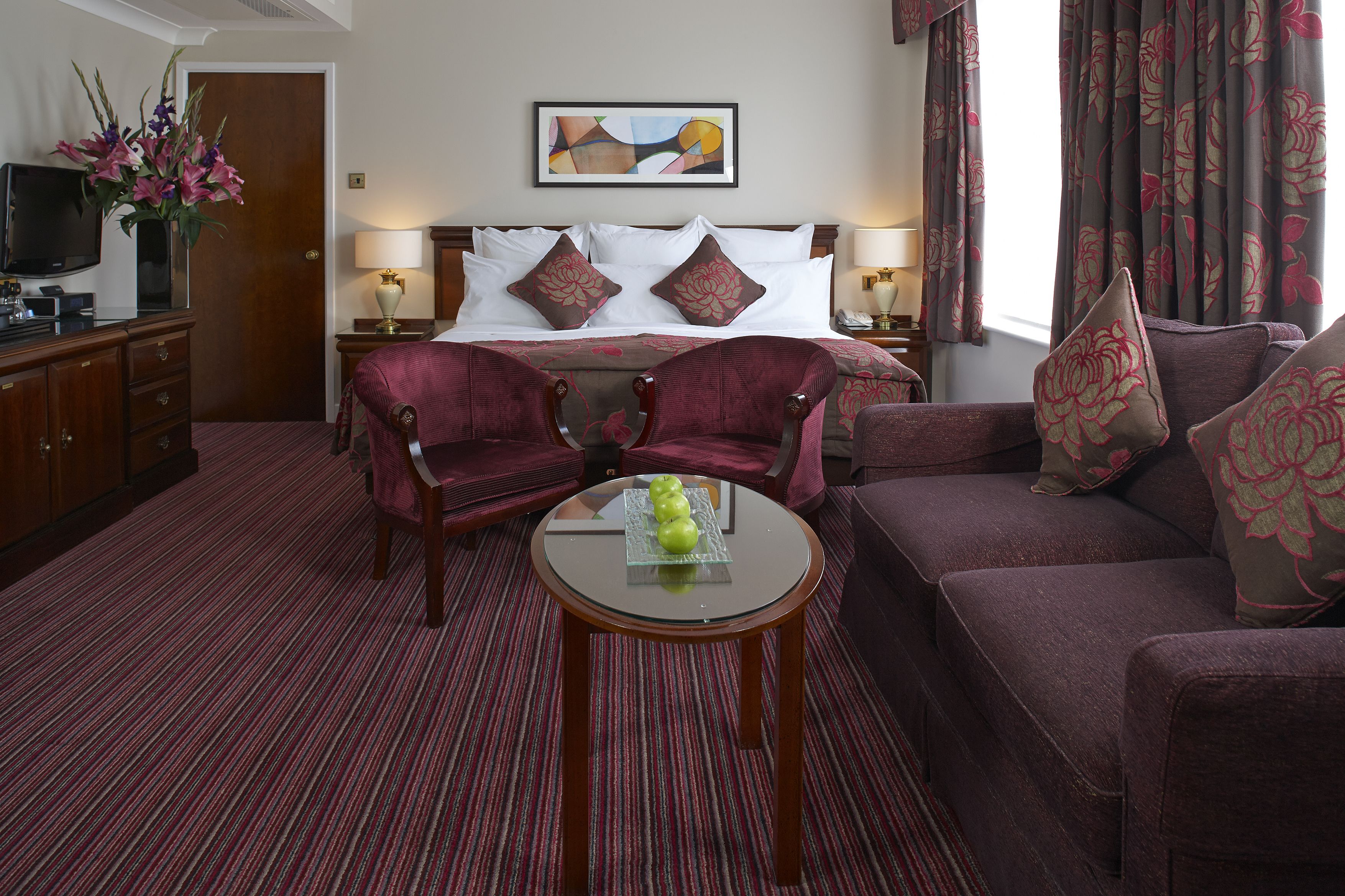 A hotel room with a bed, two armchairs, a sofa, a small table with apples, a TV, a lamp, a painting, and curtains.