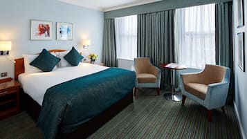 A hotel room with a large bed, two chairs, a small table, and artwork on the wall.