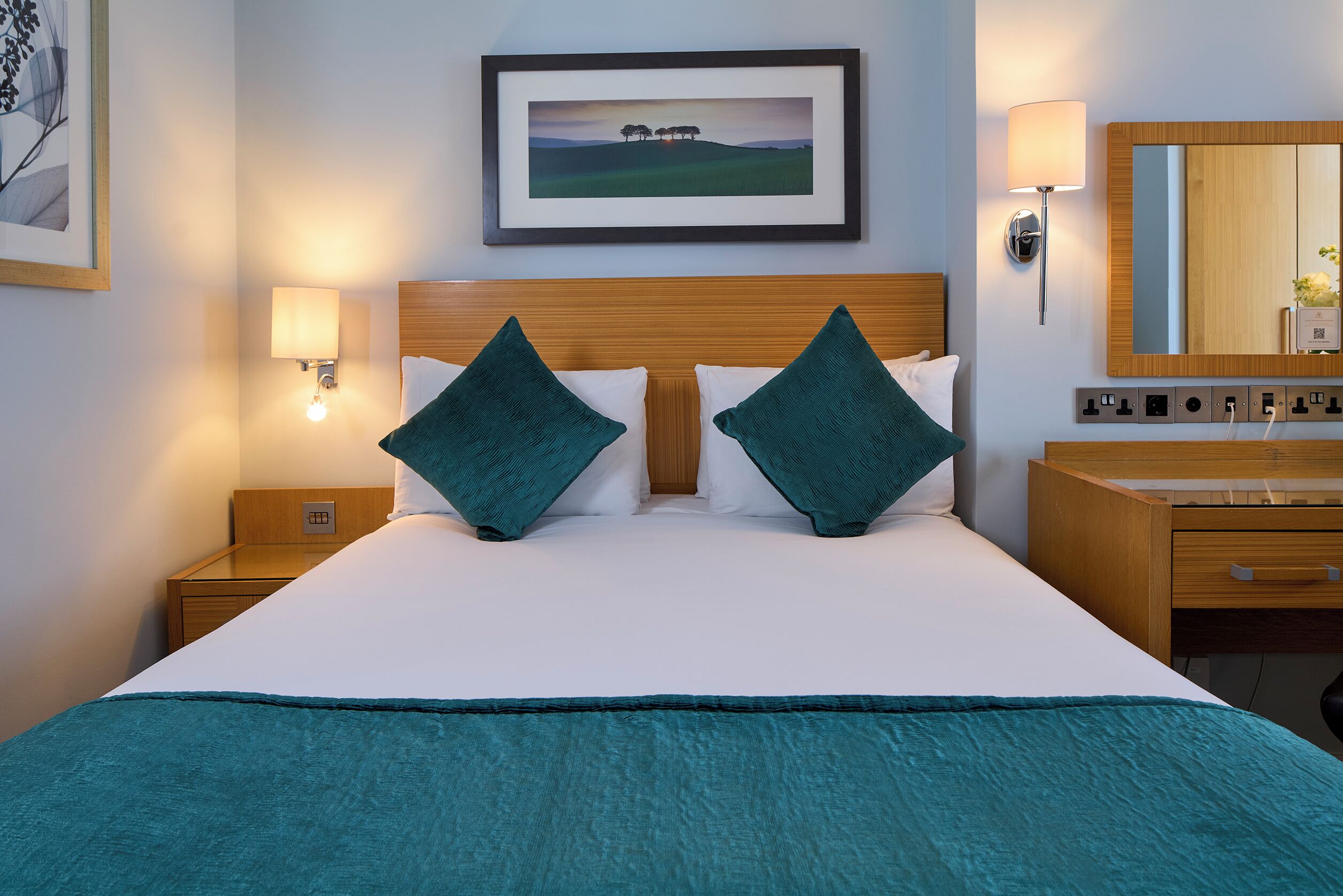 A neatly made bed with teal bedding, a wooden headboard, and a framed landscape painting above it.