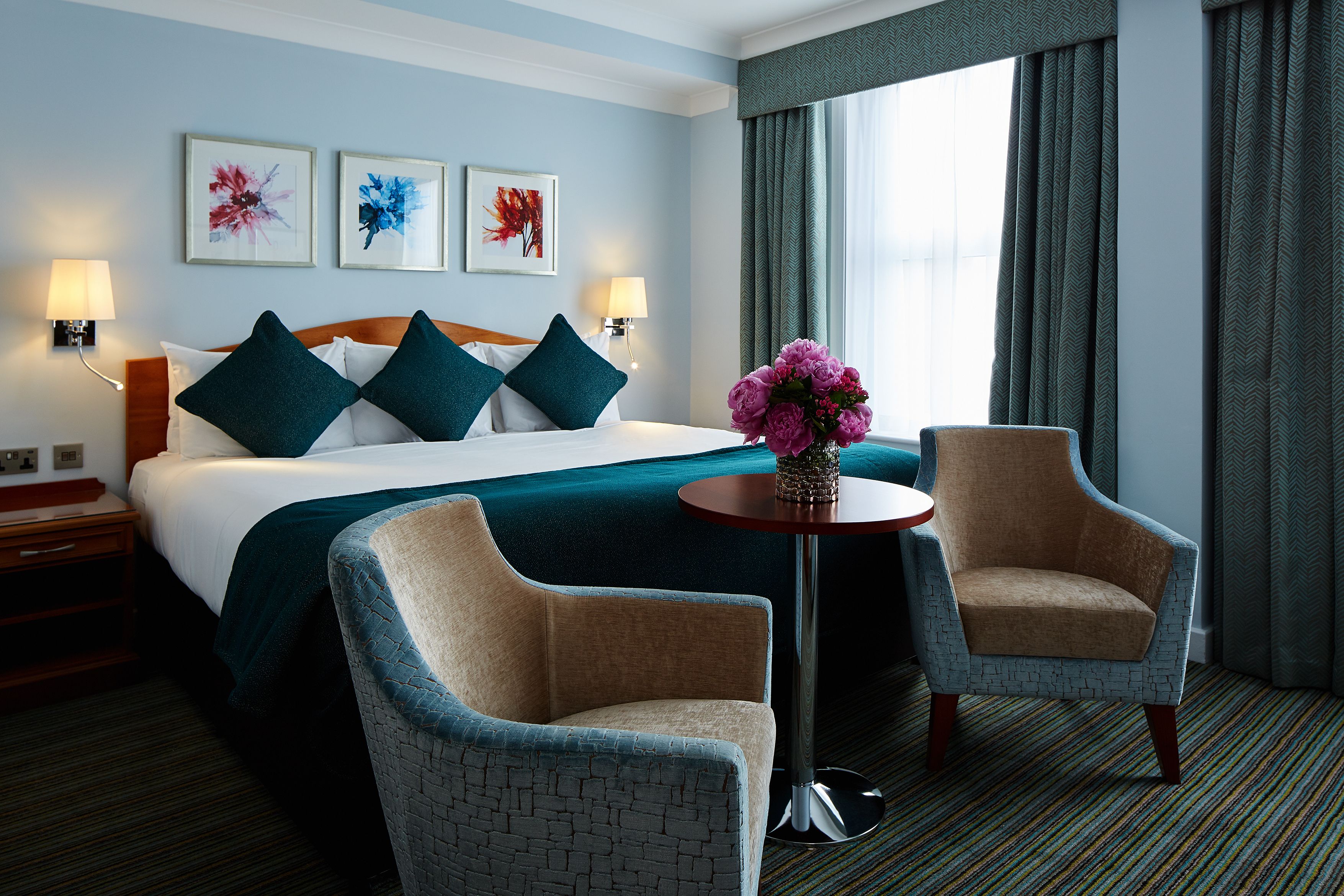 A hotel room with a large bed, two armchairs, a small table, and artwork on the wall.