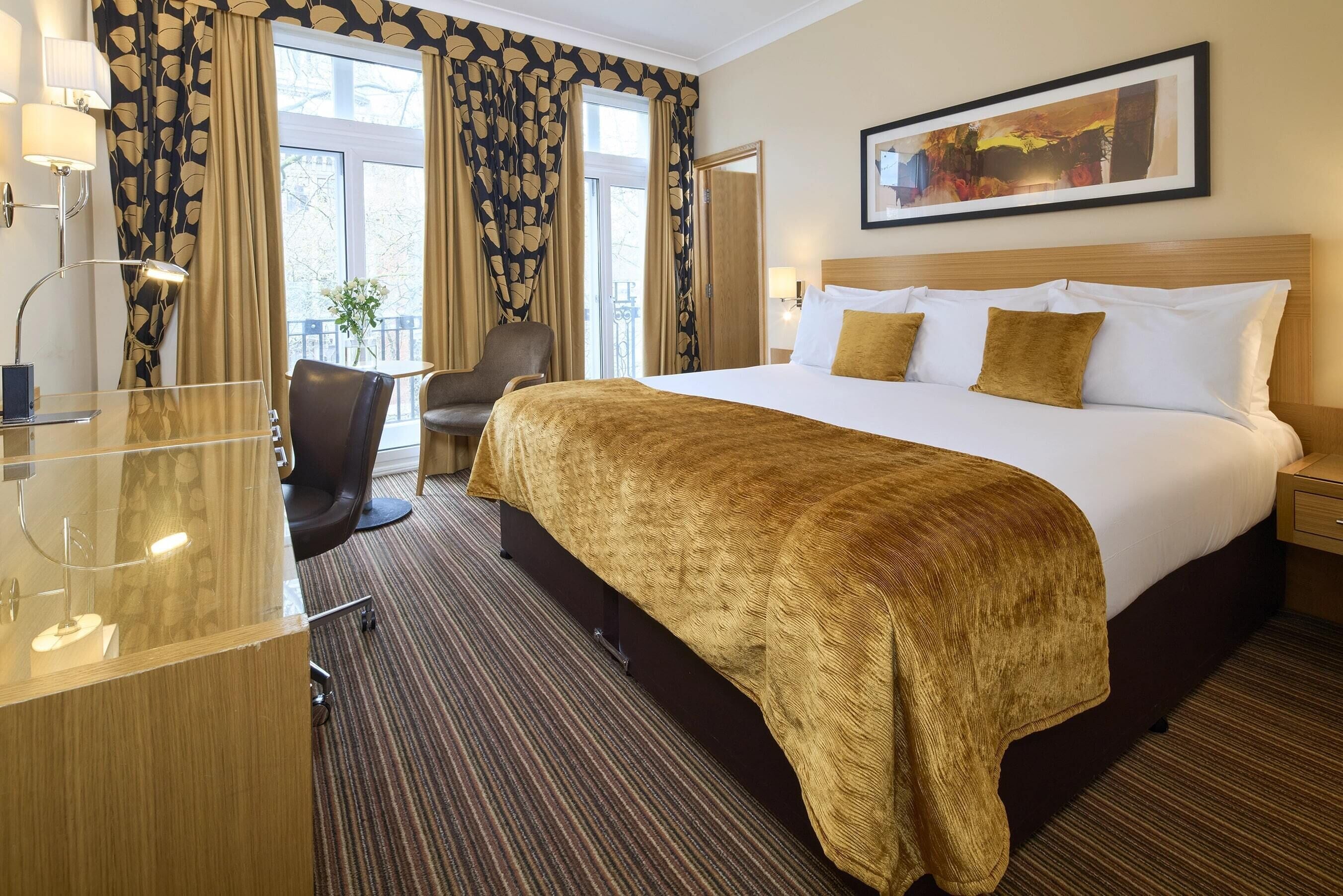 Une chambre d’hôtel avec un grand lit, un bureau avec une lampe, une chaise et une fenêtre avec des rideaux.