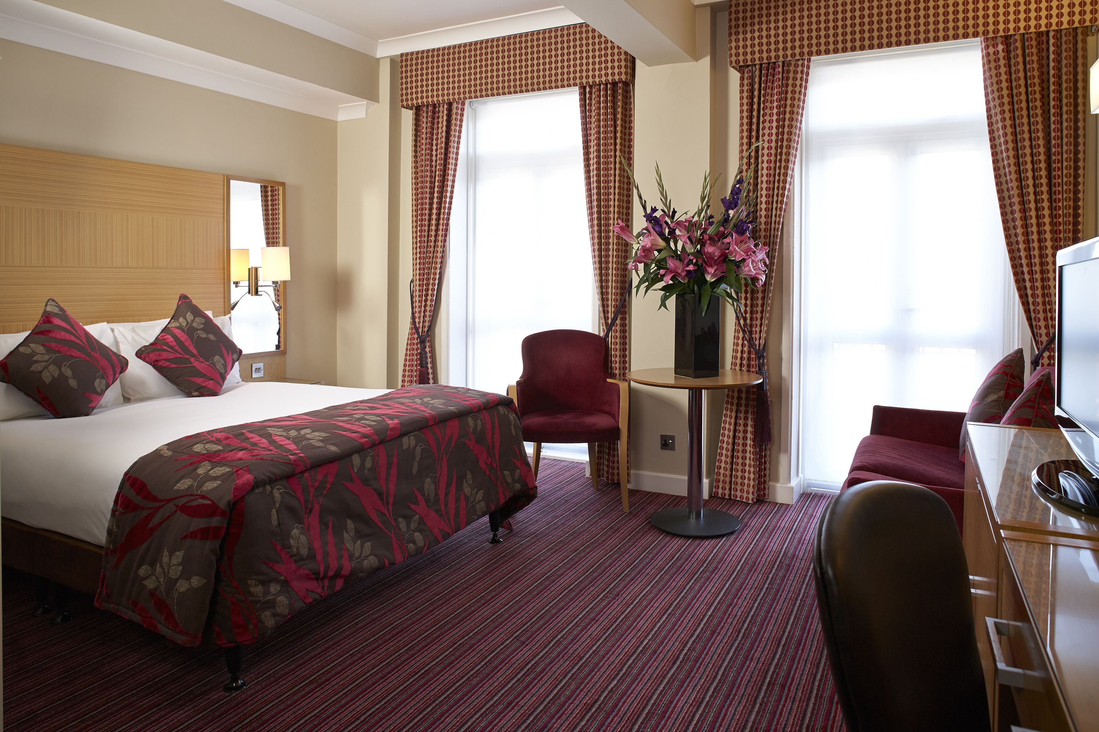 Quarto de hotel com uma cama grande, uma mesa de trabalho, uma cadeira e uma pequena mesa com um vaso de flores.