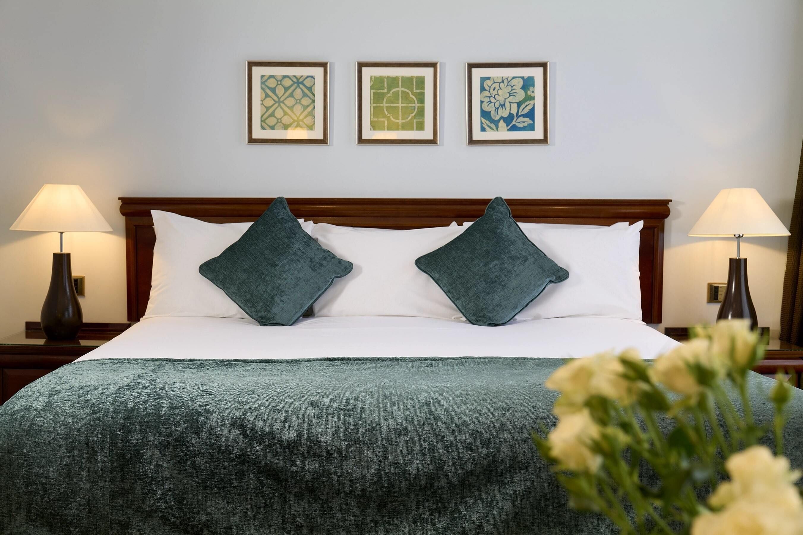 A neatly made bed with a green blanket and two pillows, flanked by two bedside lamps and framed artwork on the wall.
