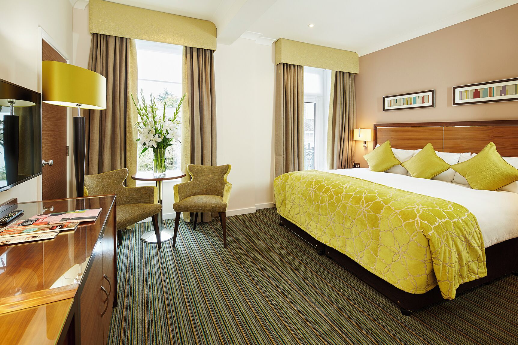 A hotel room with a bed, a desk, two chairs, a TV, and a window with curtains.
