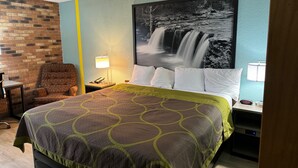 A hotel room with a bed, a chair, a nightstand, a lamp, and a wall with a waterfall painting.