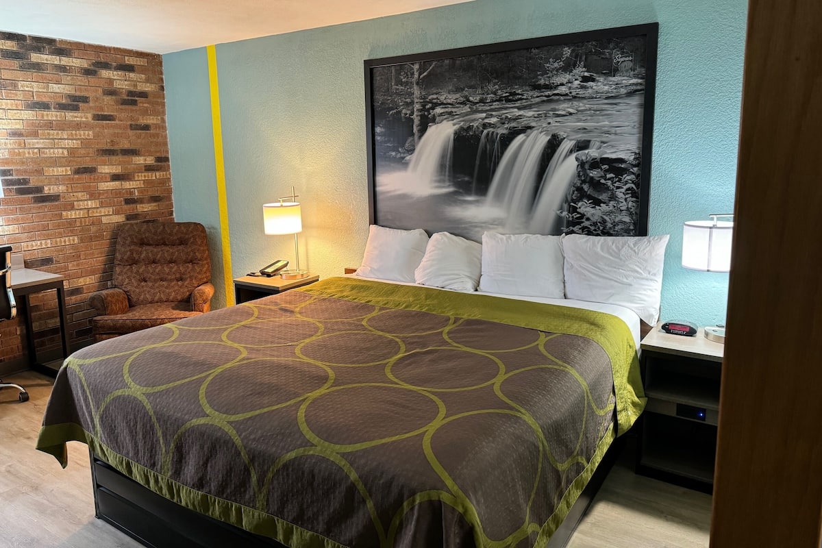 Ein Hotelzimmer mit Bett, Sessel, Nachttisch, Lampe und Wandbild mit einem Wasserfall.