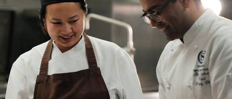 Duas pessoas em uma cozinha, uma vestindo chapéu e avental de chef, e a outra com uniforme branco de chef, com uma bandeja de pequenos recipientes na bancada.