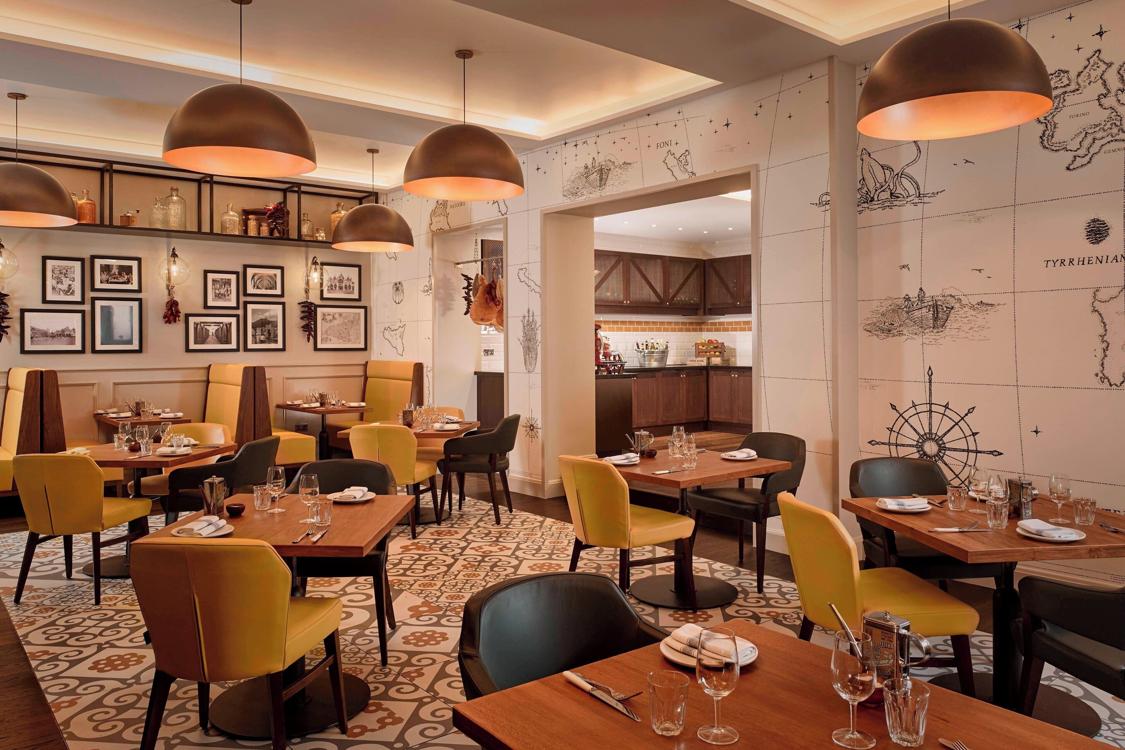 A restaurant with multiple dining tables and chairs, overhead pendant lighting, and a wall decorated with framed pictures and a map.