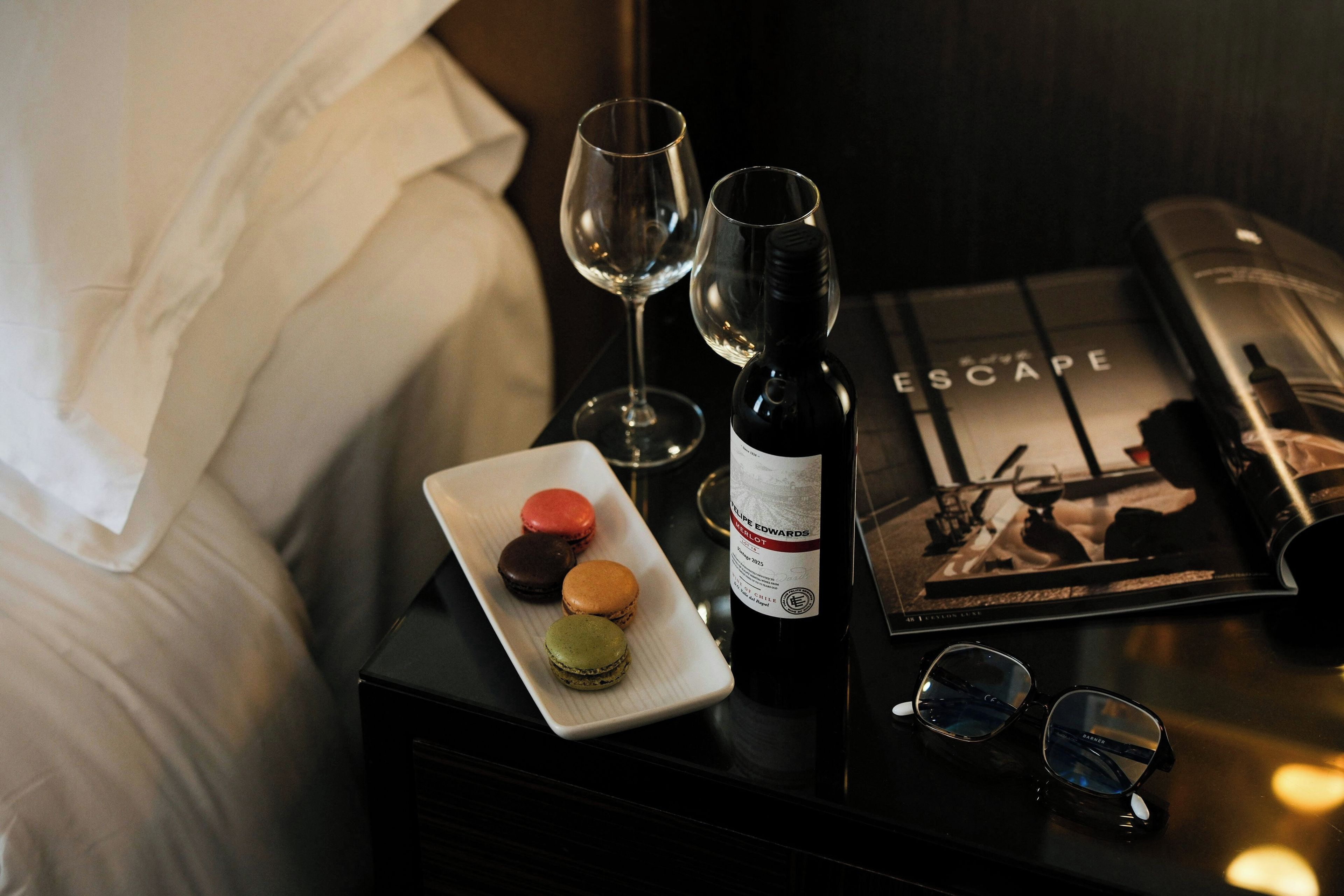 A neatly arranged hotel room with a bed, a bedside table with a wine bottle, glasses, and a plate of macarons.