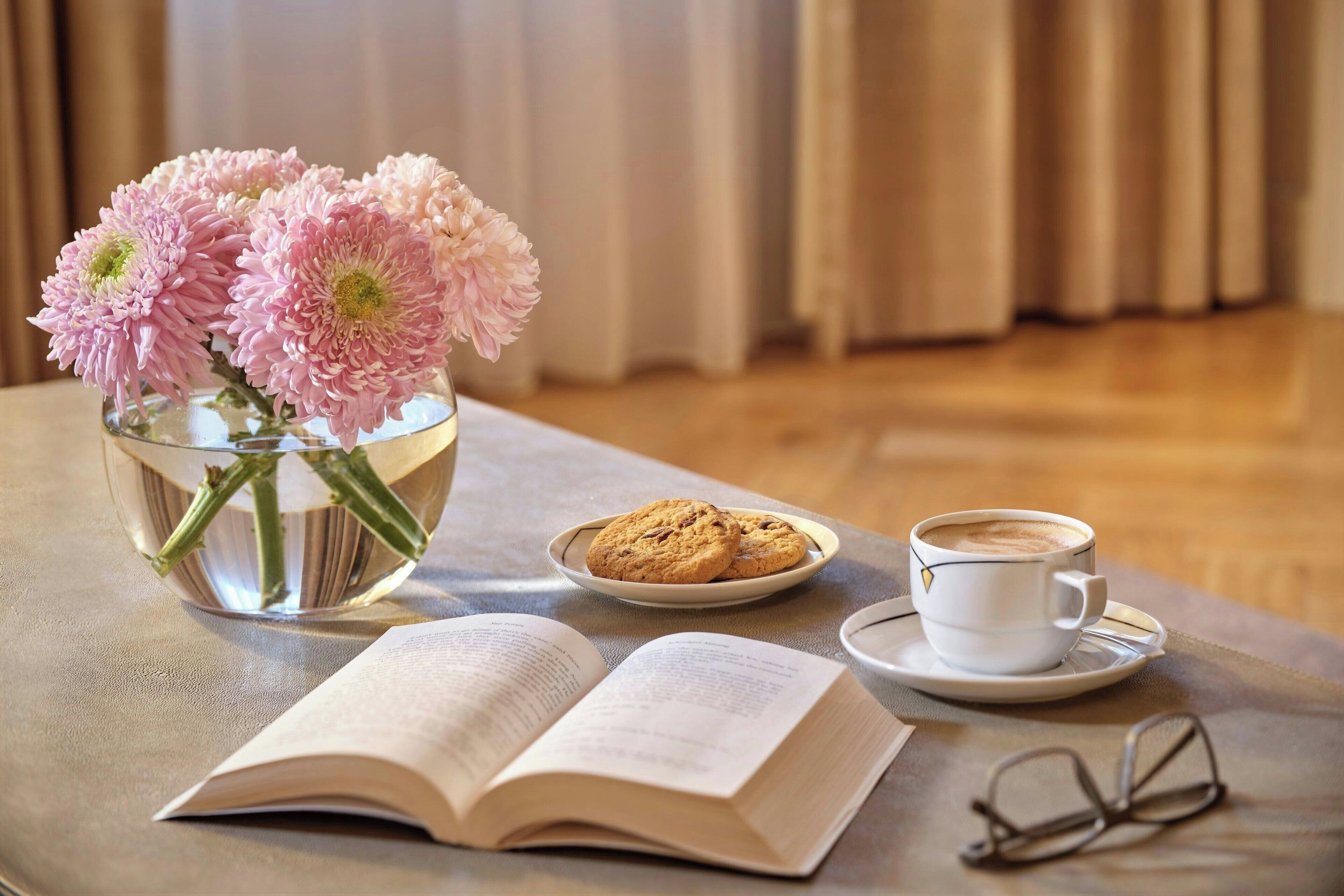 A table with a book, a cup of coffee, a plate of cookies, and a vase with flowers.