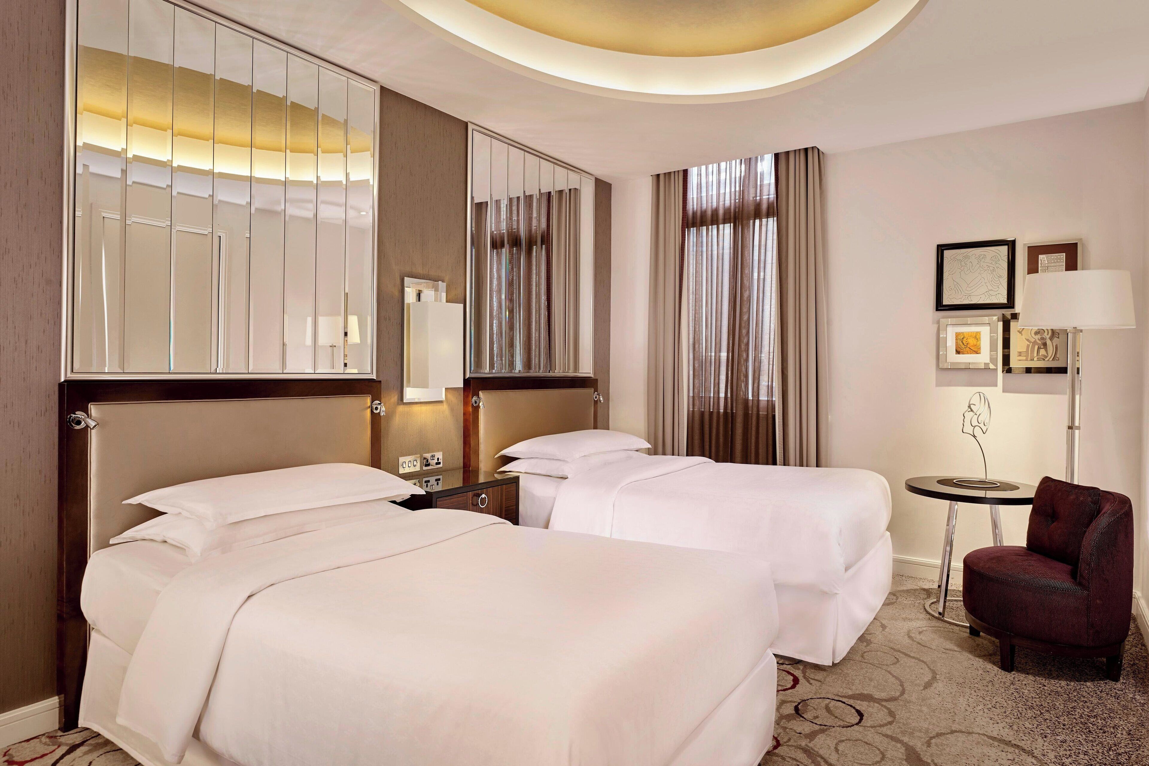 A hotel room with two beds, a small table, a chair, a lamp, and a mirror.