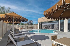 A pool area with lounge chairs and umbrellas, a pool, and a building with large windows.
