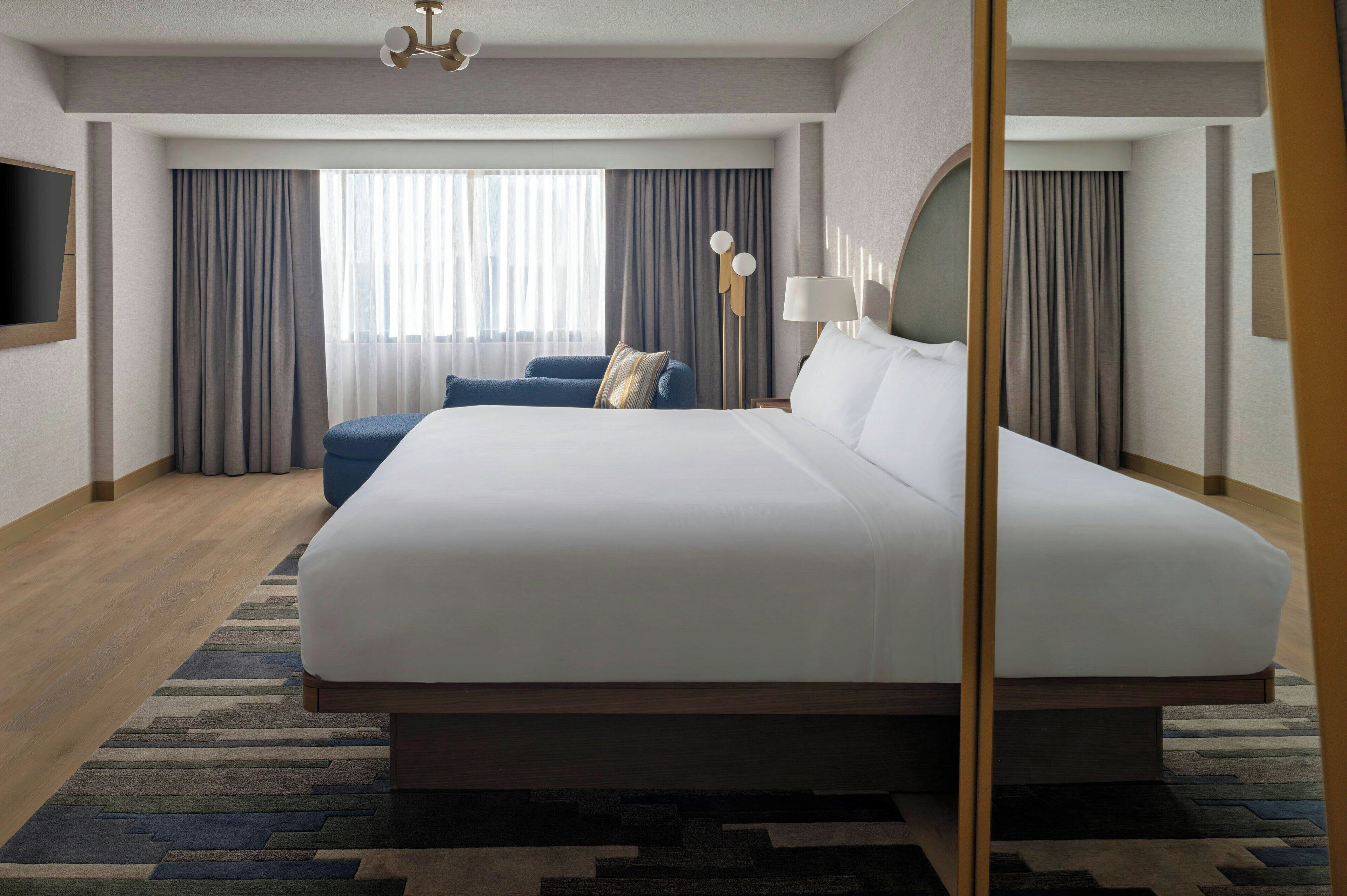 Une chambre d’hôtel moderne avec un grand lit, un fauteuil bleu et un téléviseur à écran plat fixé au mur.