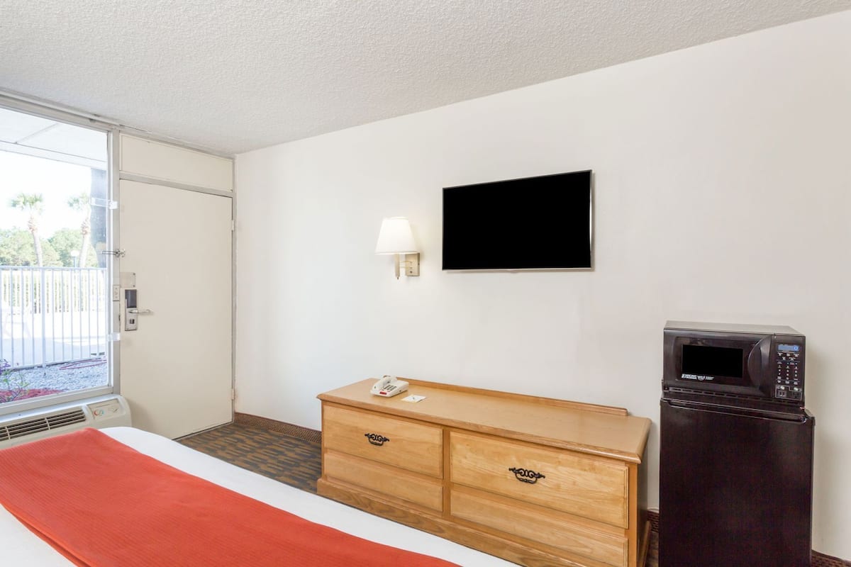 A hotel room with a bed, a wooden dresser, a television, a microwave, and a window with a view of a fence and trees.