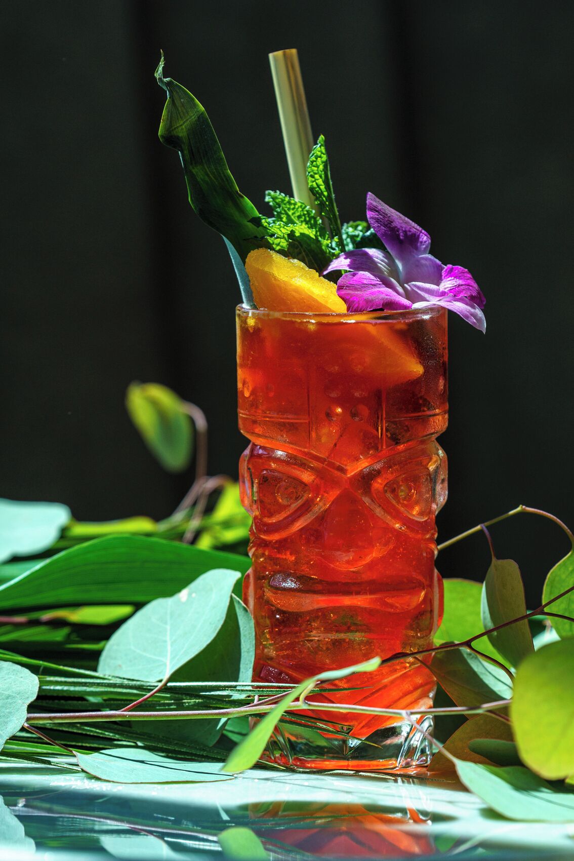 A tall glass with a textured design, filled with a red liquid, garnished with a slice of orange, purple flowers, and green leaves.