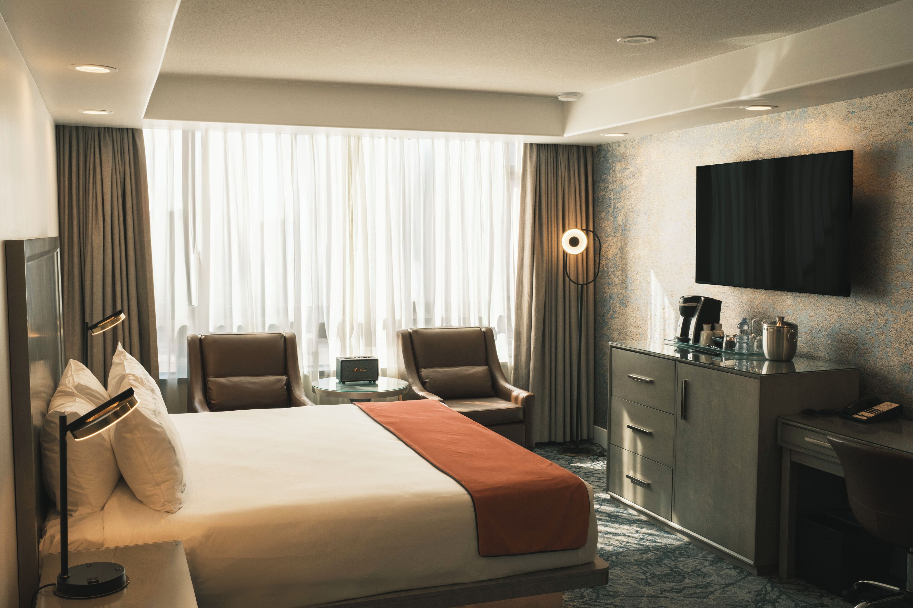 Ein Hotelzimmer mit einem Bett, zwei Stühlen, einem Schreibtisch, einem Fernseher und einem fenster mit Vorhängen.
