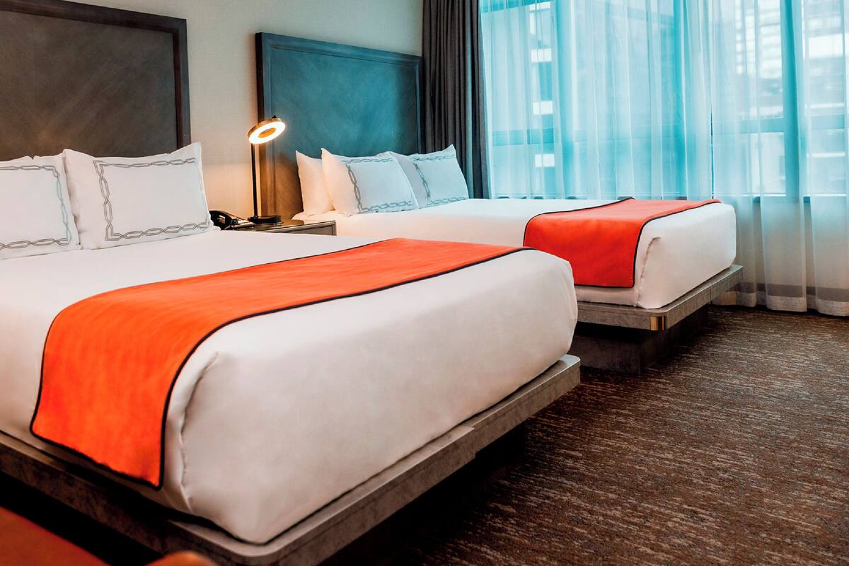 A hotel room with two beds, a nightstand, a lamp, and a view of the city through sheer curtains.
