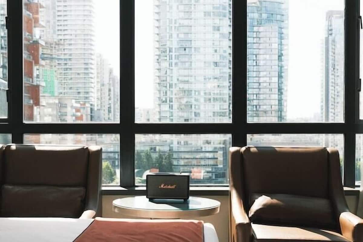 A hotel room with a bed, two armchairs, a small table, and a view of tall buildings through large windows.