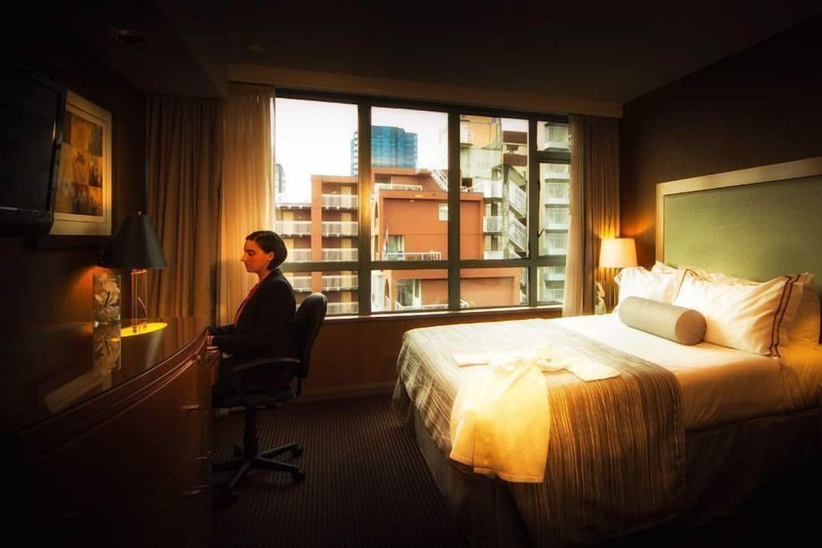 A hotel room with a large bed, a desk, a chair, and a view of the cityscape through the window.