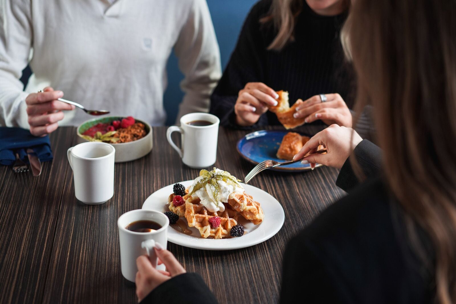 Un grupo de personas cenando en una mesa con varios platos, incluyendo waffles con frutos rojos y crema batida, y café.