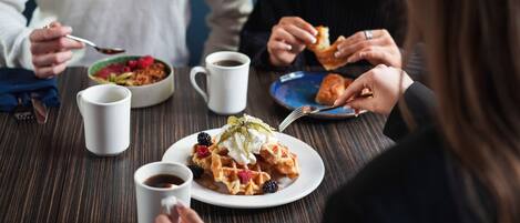 Un grupo de personas cenando en una mesa con varios platos, incluyendo waffles con frutos rojos y crema batida, y café.