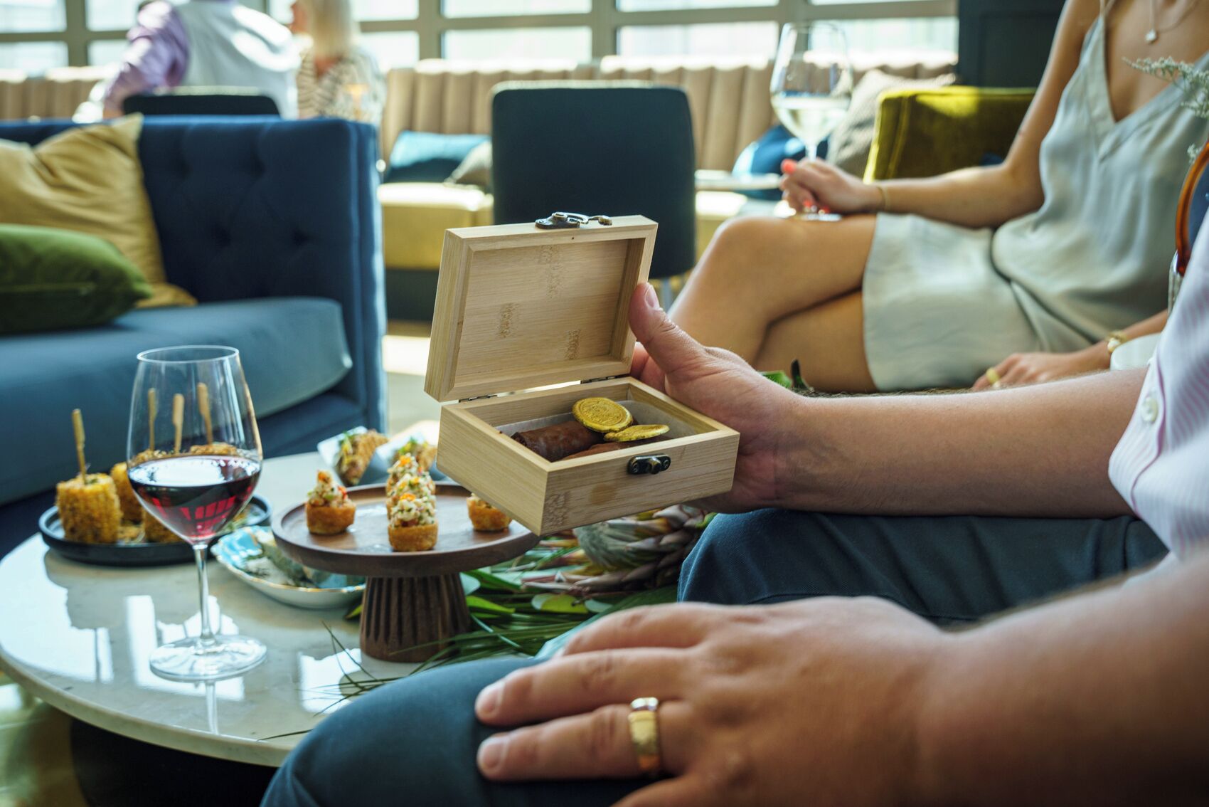 Una mesa de comedor con una caja de madera que contiene alimentos, una copa de vino y un plato de aperitivos.