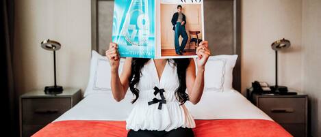 A person holding two magazines, one titled "LOVE" and the other "NUVO", in front of a bed with a red blanket and white pillows.