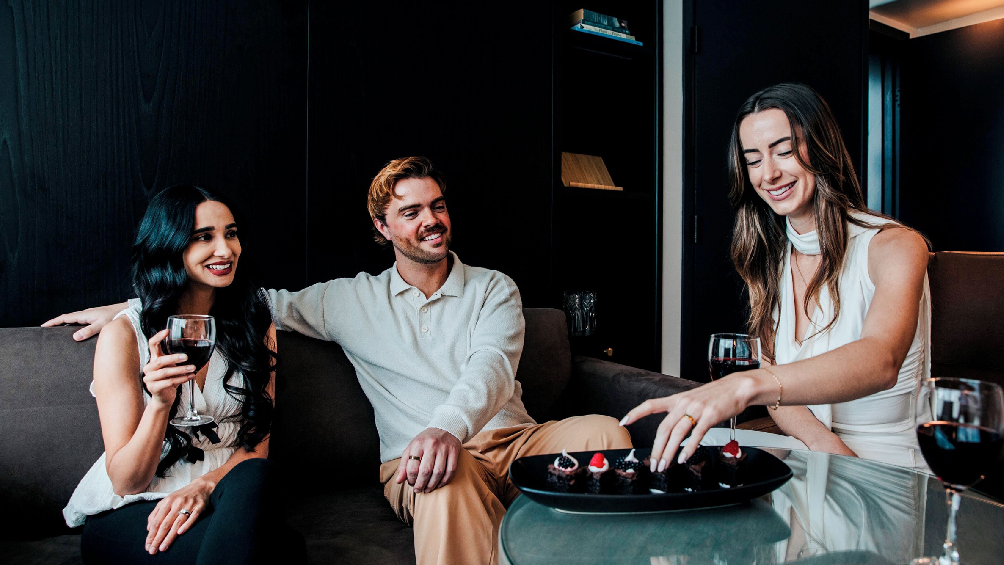 Three people sitting on a couch with a plate of desserts and wine glasses on a table.
