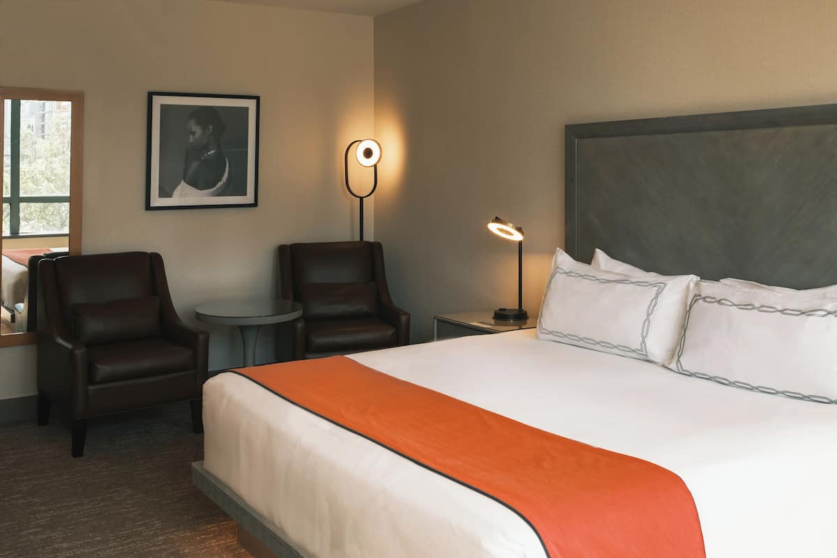 A hotel room with a large bed, two chairs, a small table, and a nightstand with a lamp.