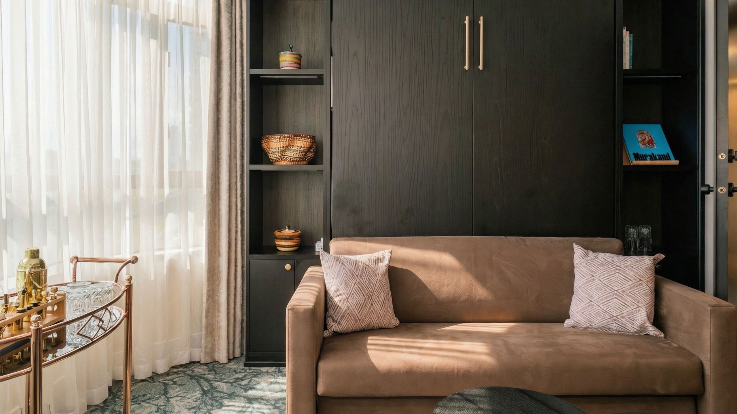 A modern living room with a beige sofa, patterned cushions, a coffee table with decorative items, and a bookshelf with books and a small blue box.