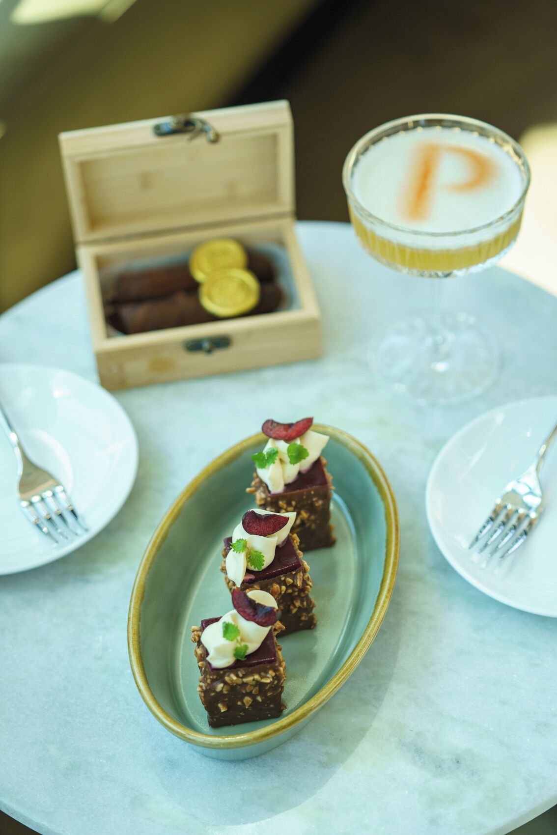 A dessert plate with three square pieces topped with cream and berries, a glass of a beverage with a frothy top, and a wooden box with chocolates in the background.